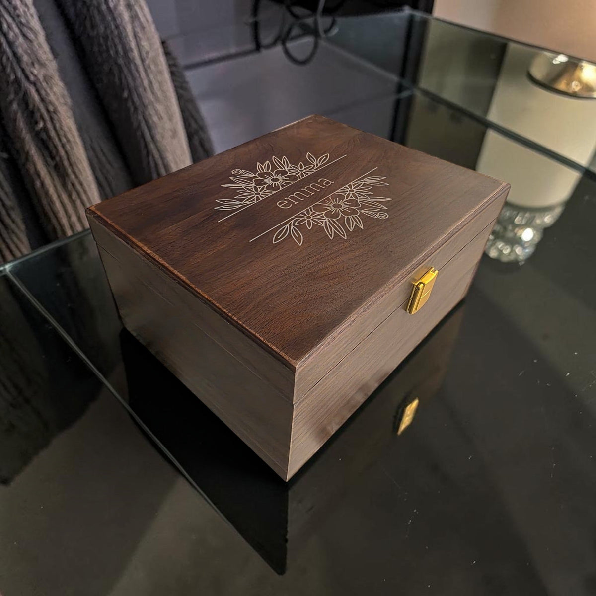 Newfashioned Relics dark wood personalized keepsake box with floral engraving and gold latch on a glass table.
